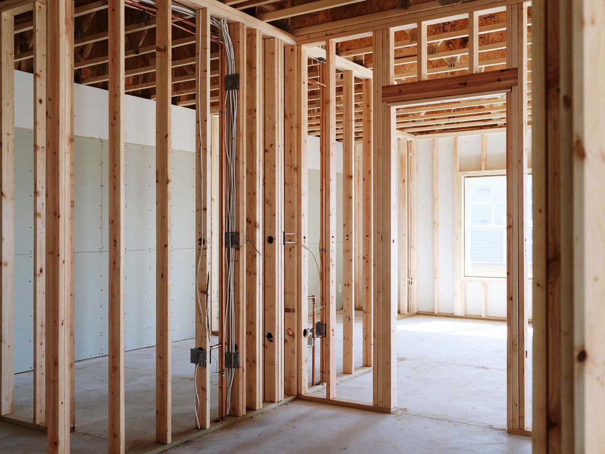 Bare wood studs of an unfinished wall with electrical cable and copper pipe run through them and protected by steel plates, a stack of pale gray sheets lying flat on the floor beyond