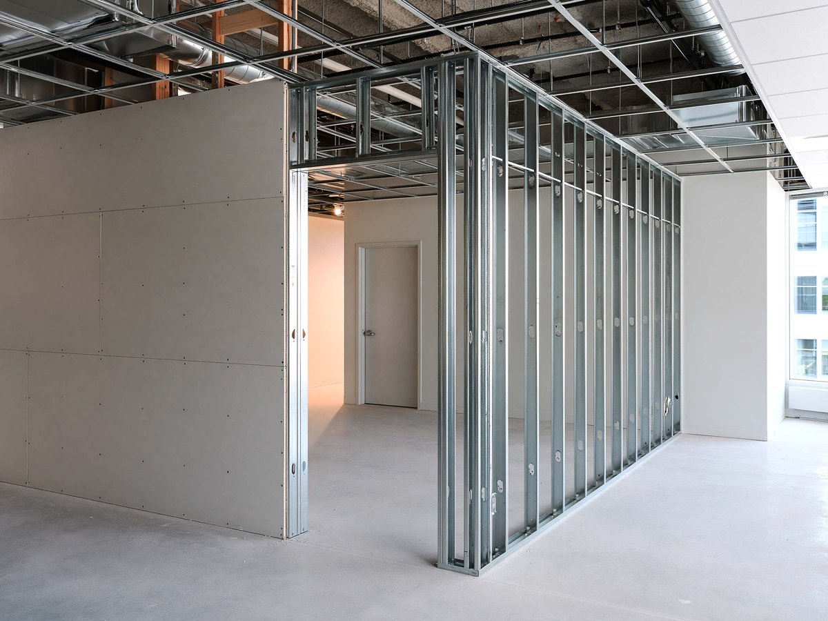 New metal stud partition inside an unfinished office suite with pale gray board hung across half of it and the framing still open on the other half, a rolled set of drawings on a folding table in the foreground