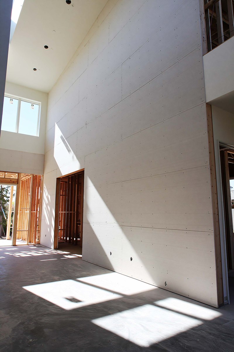Interior of a house under construction with pale gray drywall board hung across the studs of two rooms and a hallway, daylight through an unglazed opening, the floor bare and swept